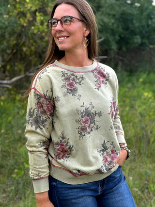A woman wearing a sage green crewneck sweatshirt with a vintage floral print and pinkish-red trim, paired with blue jeans, standing outdoors in a grassy area.