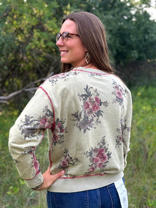 A woman wearing a cream-colored crewneck sweatshirt with a dusty rose and gray floral print, photographed from behind in an outdoor setting with green foliage in the background.