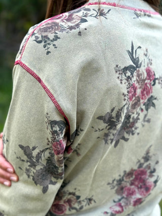 Close-up of a person wearing a light beige or sage green long-sleeve top with a dark floral print and pink stitching along the shoulder seams.
