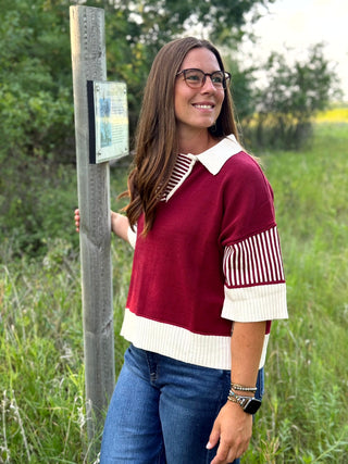 A woman wearing a burgundy and cream knit sweater with striped details stands outdoors next to a wooden signpost in a grassy field.