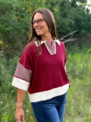 A woman wearing a burgundy knit top with cream ribbed trim and striped sleeve panels, paired with blue jeans, standing outdoors in a grassy area.
