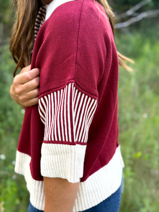 A close-up of a person wearing a maroon knit cardigan with cream ribbed trim and a cream-and-maroon striped sleeve panel, photographed outdoors with blurred greenery in the background.