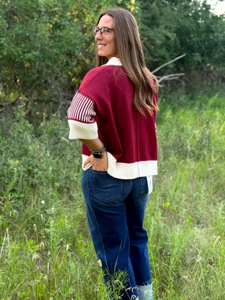 A woman wearing a burgundy and cream knit cardigan with striped sleeves, blue jeans, and glasses, standing outdoors in a grassy field.