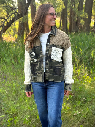 A woman wearing a mixed-media patchwork jacket with quilted tan sections, camouflage print panels, and cream cable-knit sleeves, standing outdoors in a grassy wooded area.