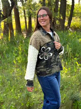 A woman wearing a mixed-media quilted jacket with a brown quilted yoke, white cable-knit sleeves, and a black camouflage vest body with a white floral applique, standing outdoors in a grassy wooded area.