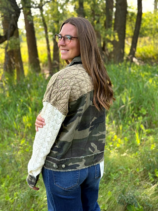 A woman wearing a mixed-media jacket with quilted tan shoulders, a camouflage body, and a cream cable-knit sleeve, standing in a grassy wooded area.