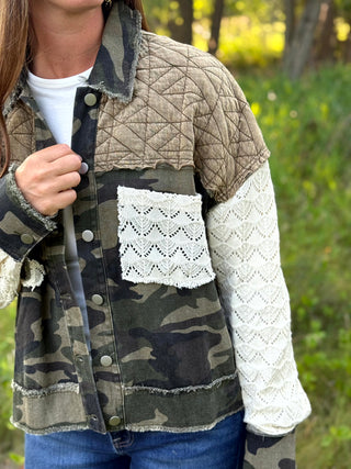 A person wearing a mixed-media patchwork jacket outdoors. The jacket features a quilted tan upper section, a green and brown camouflage lower section, a white lace pocket, and a white textured knit sleeve.