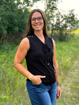 A woman wearing a sleeveless black blouse with a tie-neck detail and blue jeans, standing outdoors in a grassy field.