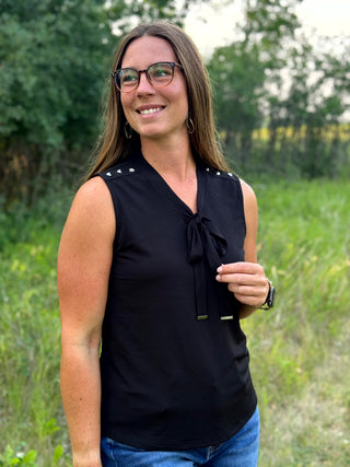 A woman wearing a black sleeveless blouse with a tie-neck detail and small silver studs on the shoulders, standing outdoors in a grassy field.
