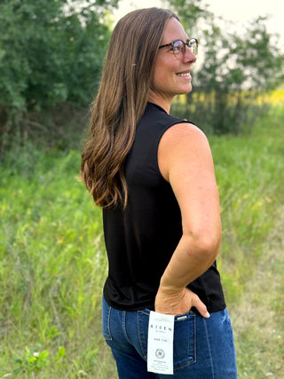 A woman wearing a sleeveless black top and blue jeans stands outdoors in a grassy field, looking back over her shoulder. A white hangtag is visible at the back of the jeans.