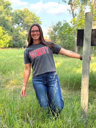 A woman standing in a grassy field outdoors, wearing a dark gray graphic t-shirt with 'HOWDY' printed in red and blue, paired with blue jeans.