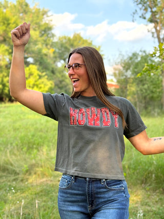A woman outdoors wearing a faded charcoal gray crop top t-shirt with 'HOWDY' printed in distressed red letters across the chest, paired with blue jeans.