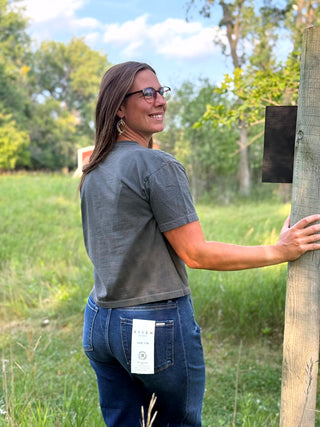 A woman wearing a dark gray t-shirt and blue jeans stands outdoors next to a wooden post, looking back over her shoulder. A white tag is visible on the back pocket of her jeans.