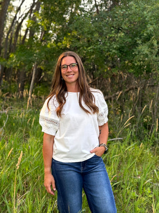 A woman with glasses stands outdoors in a grassy wooded area, wearing a white short-sleeve top with lace puff sleeves and blue jeans.