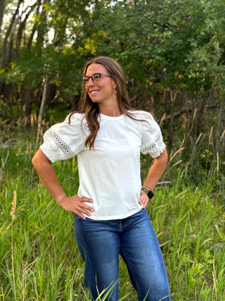 A woman wearing a white short-sleeve blouse with lace puff sleeves and blue jeans, standing outdoors in a grassy field with trees in the background.
