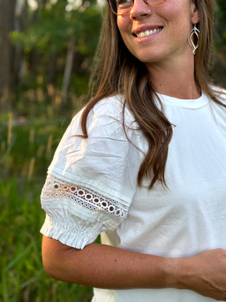 A woman wearing a white short-sleeve blouse with eyelet lace detailing on the sleeve, photographed outdoors against a blurred green background.