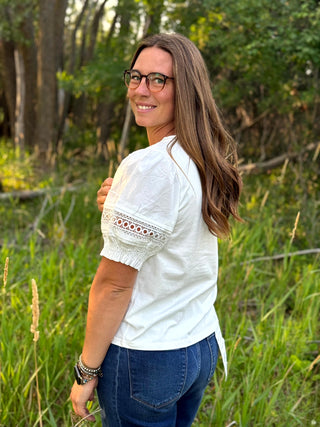 A woman with long brown hair and glasses, wearing a white short-sleeve blouse with embroidered puff sleeves and blue jeans, standing in a grassy outdoor setting.