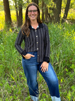 A woman wearing a black and white vertically striped button-up shirt with a dark collar and cuffs, paired with blue jeans, standing outdoors in a grassy wooded area.