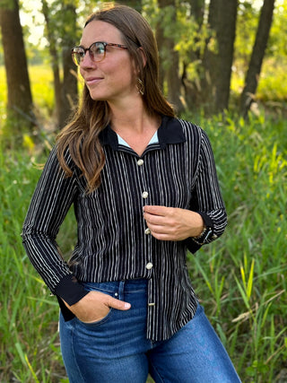 A woman wearing a black and white vertically striped button-up shirt with a solid black collar, standing outdoors in a grassy wooded area.