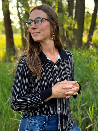 A woman wearing a black and white vertically striped button-up shirt with a solid black collar, standing outdoors in a grassy area with trees in the background.