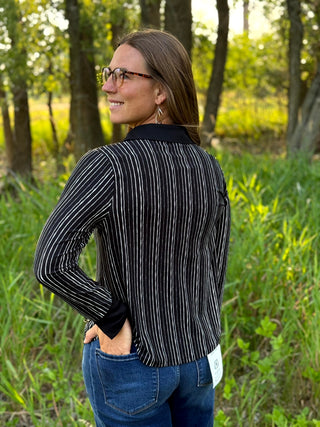 A woman wearing a black and white vertically striped long-sleeve shirt and blue jeans, photographed outdoors in a grassy wooded area.