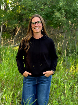 A woman wearing a black pullover hoodie with drawstrings and gold-toned hardware, paired with blue jeans, standing outdoors in a grassy field with trees in the background.
