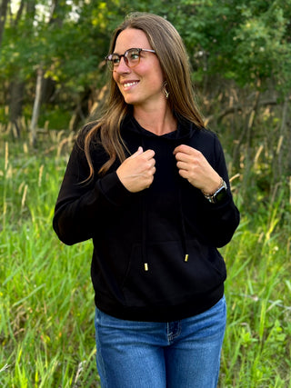 A woman wearing a black pullover hoodie with drawstrings and gold-toned hardware, paired with blue jeans, standing outdoors in a grassy field with trees in the background.