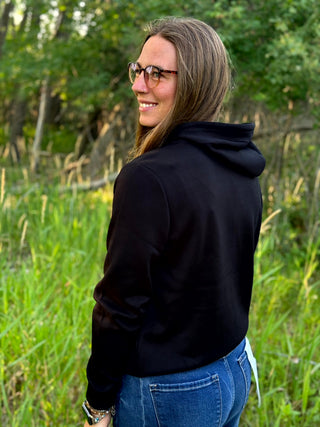 A woman with glasses is shown from the back wearing a black cropped hoodie and blue jeans, standing outdoors in a grassy field with trees in the background.