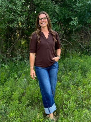 A woman wearing a brown short-sleeve henley shirt and blue jeans stands outdoors in a grassy area with trees in the background.