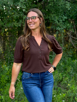 A woman wearing a brown short-sleeve henley top and blue jeans, standing outdoors in front of a green forest background.