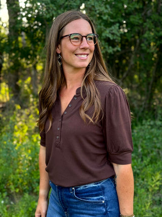 A woman with long brown hair and glasses wearing a dark brown ribbed short-sleeve henley top and blue jeans, standing outdoors in front of a blurred green forest background.