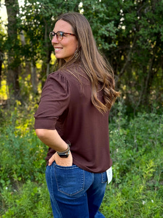 A woman with long brown hair and glasses, wearing a brown short-sleeve top and blue jeans, standing outdoors in a grassy, wooded area.