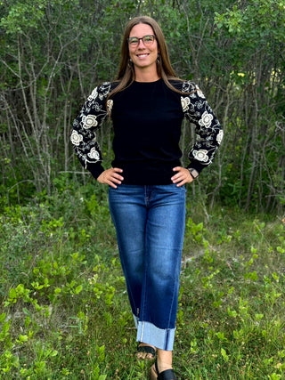 A woman wearing a black long-sleeve sweater with white floral embroidered sleeves, blue jeans, and glasses, standing outdoors in a grassy, wooded area.