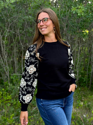 A woman wearing a black crewneck sweater with white floral embroidery on the sleeves, paired with blue jeans, standing outdoors in front of a green forest background.