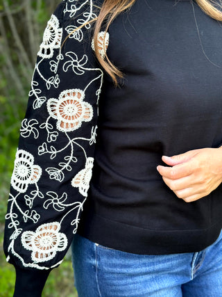 A close-up of a person wearing a black long-sleeve top with white floral lace embroidery on one sleeve, paired with blue jeans, outdoors.