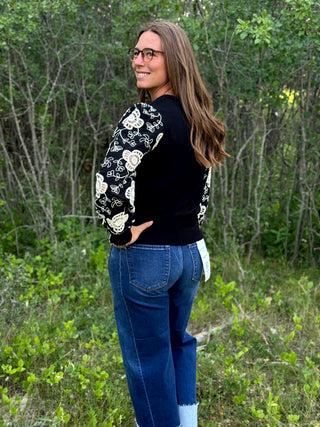 A woman wearing a black long-sleeve sweater with cream floral embroidery on the sleeves, paired with blue jeans, standing outdoors in a wooded setting.