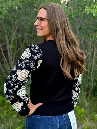 A woman wearing a black long-sleeve sweater with white floral crochet embroidery on the sleeves, photographed outdoors against a blurred green forest background.