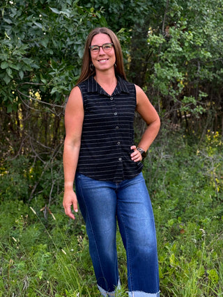 A woman wearing a sleeveless black button-front top with thin white stripes and blue jeans, standing outdoors in a grassy area with green trees in the background.
