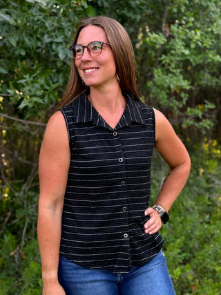 A woman wearing a sleeveless black button-front top with thin white stripes and blue jeans, standing outdoors in front of a green leafy background.