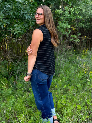 A woman wearing a black sleeveless top with thin white horizontal stripes and blue jeans, standing outdoors in a grassy area with trees in the background.