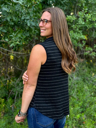 A woman with long brown hair and glasses, wearing a sleeveless black top with thin white stripes and blue jeans, standing outdoors in front of a green leafy background.