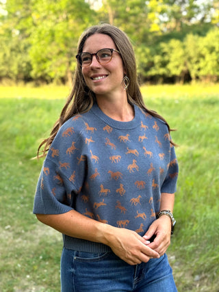 A woman wearing a slate blue crewneck sweater with an allover brown horse and rider pattern, standing outdoors in a grassy field.