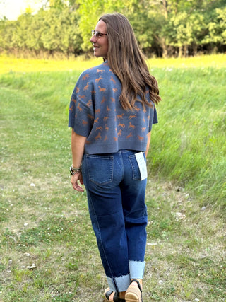 A woman standing outdoors in a grassy field, shown from the back, wearing a slate blue knit sweater with a brown animal print, dark blue jeans, and sandals.