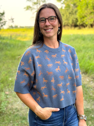 A woman wearing a slate blue short-sleeve knit sweater with an allover pattern of brown horses, standing outdoors in a grassy field.