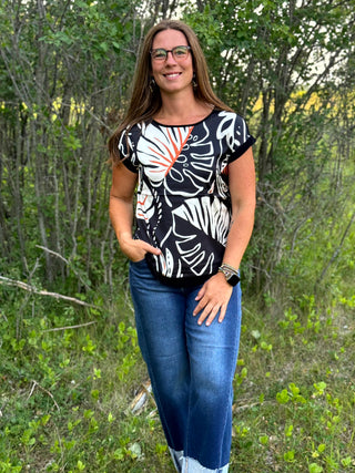 A woman wearing a black short-sleeve top with a white and orange botanical leaf pattern, paired with blue jeans, standing outdoors in a wooded setting.