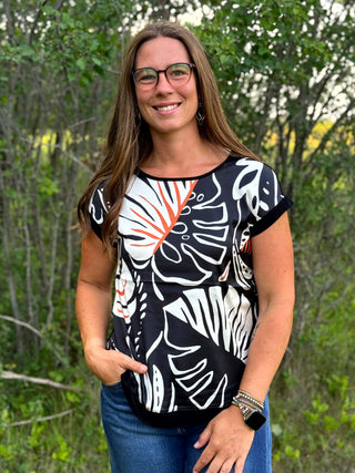 A woman wearing a black short-sleeve top with a white and orange abstract botanical pattern, paired with blue jeans, standing outdoors in front of a blurred green forest background.