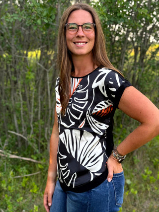 A woman wearing a black short-sleeve top with a bold white and orange botanical leaf pattern, paired with blue jeans, standing outdoors in front of a blurred green forest background.