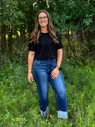 A woman wearing a black short-sleeve top and blue jeans stands outdoors in a grassy area with trees in the background.