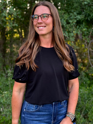 A woman with long brown hair and glasses wearing a black short-sleeve top with embellished puff sleeves and blue jeans, standing outdoors against a blurred green foliage background.