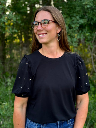 A woman with glasses and hoop earrings wearing a black short-sleeve top with ruffled sleeves and small silver stud accents, standing outdoors against a blurred green foliage background.
